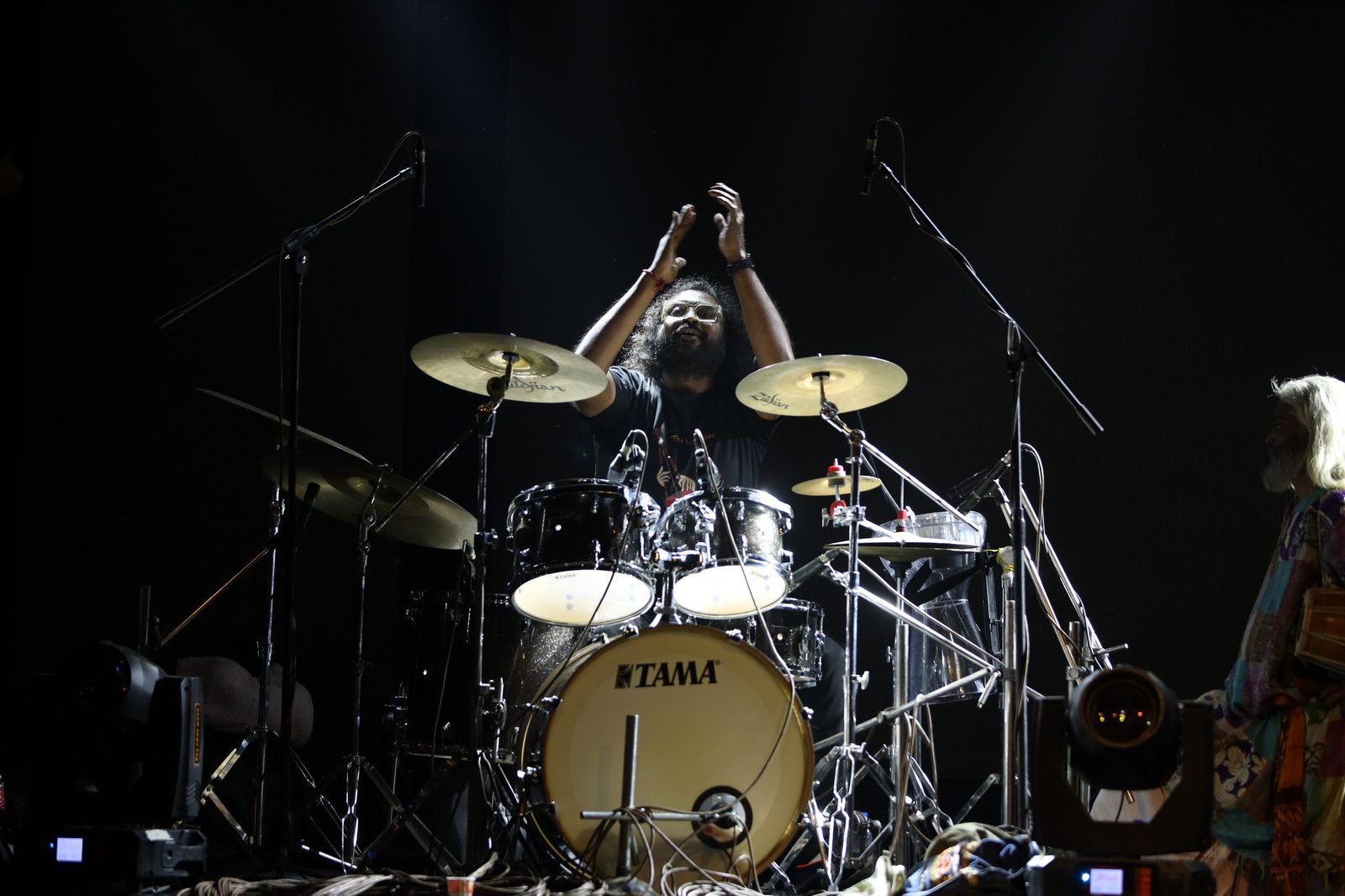 The drummer with both sticks raised above the kit, caught in a single spotlight