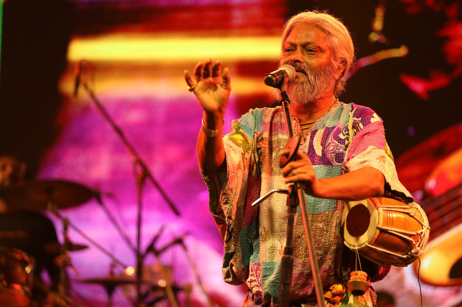 A silver-haired singer in a patchwork kantha kurta at the mic, a small hand drum slung at his hip