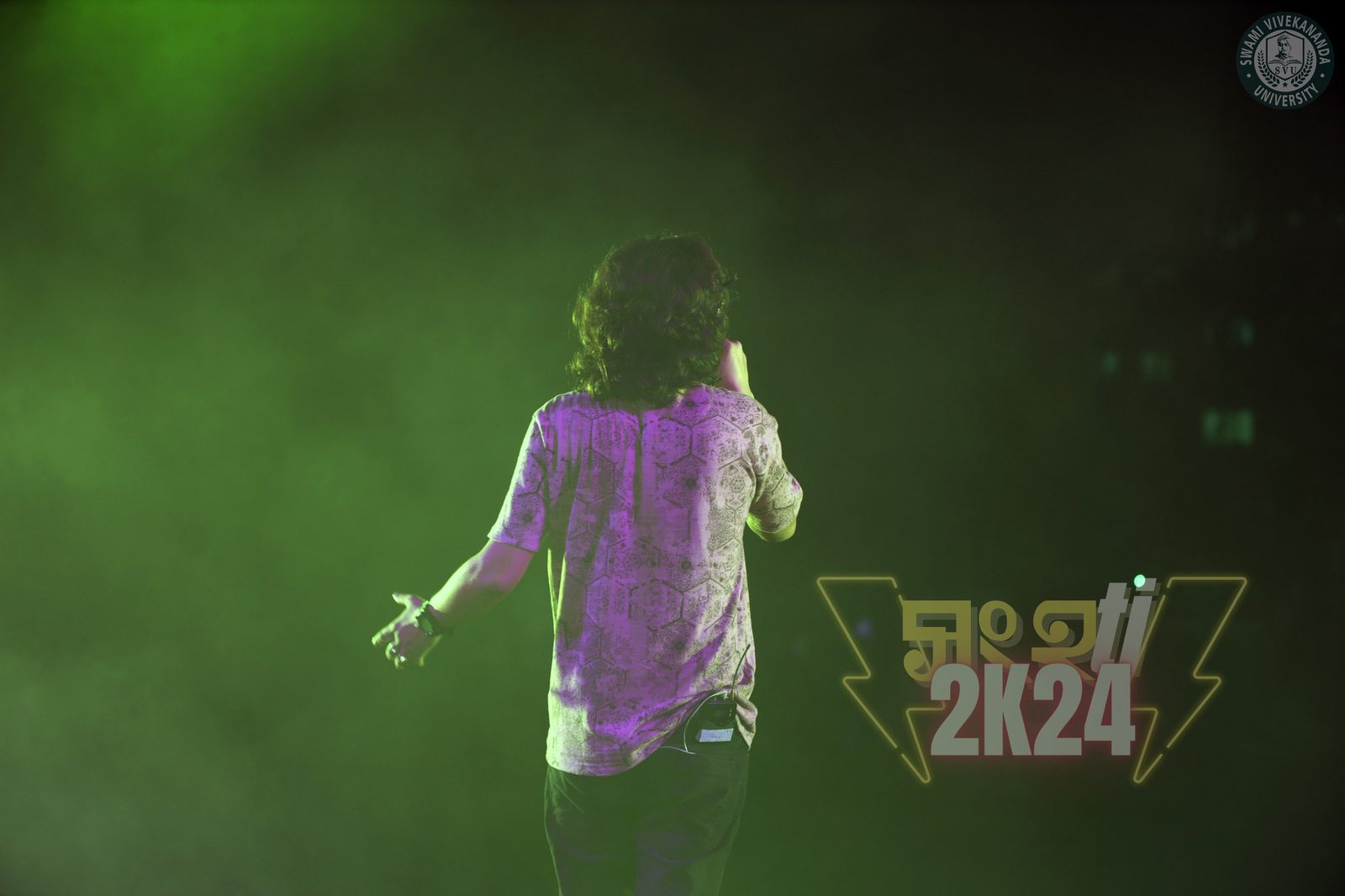 A performer from behind, arms open, the stage filled with green haze