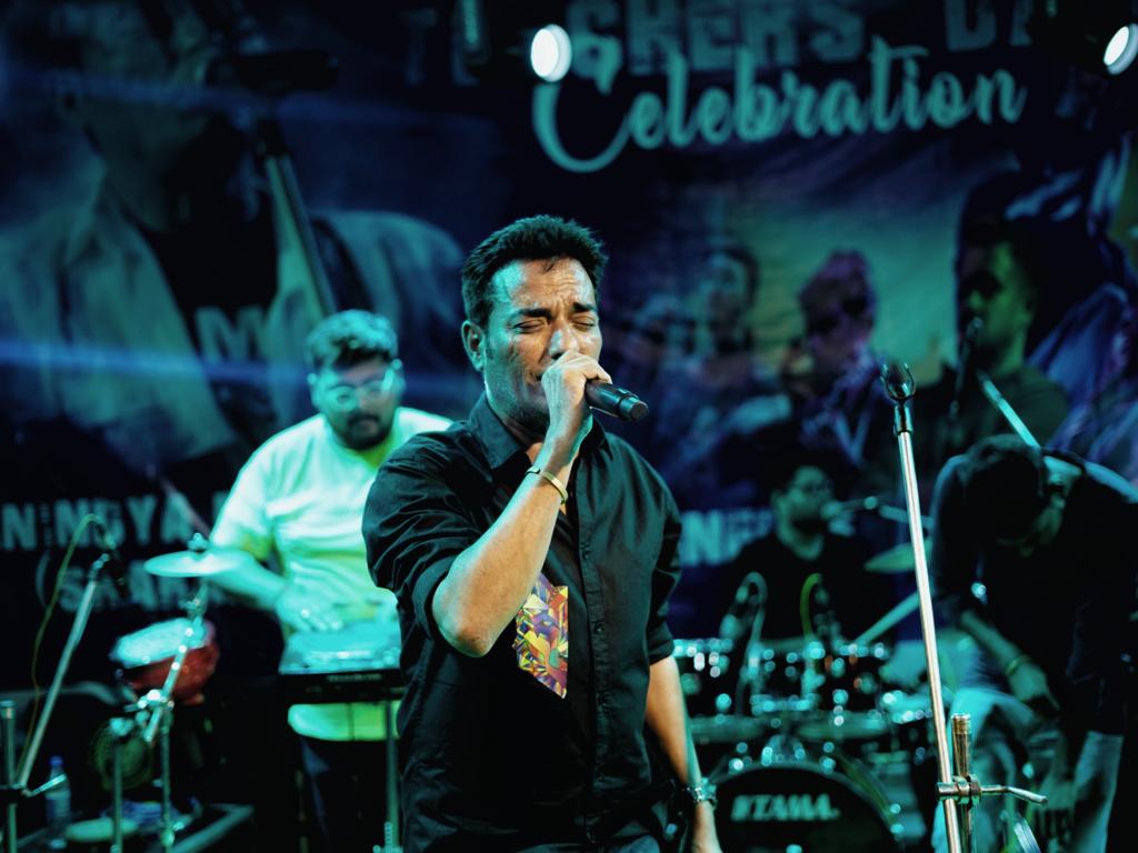 A vocalist in a black shirt singing with his eyes shut, the band lit blue behind him