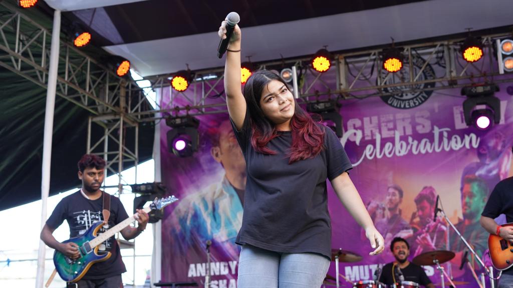 A singer holding her microphone above her head, guitarist and drummer playing behind her