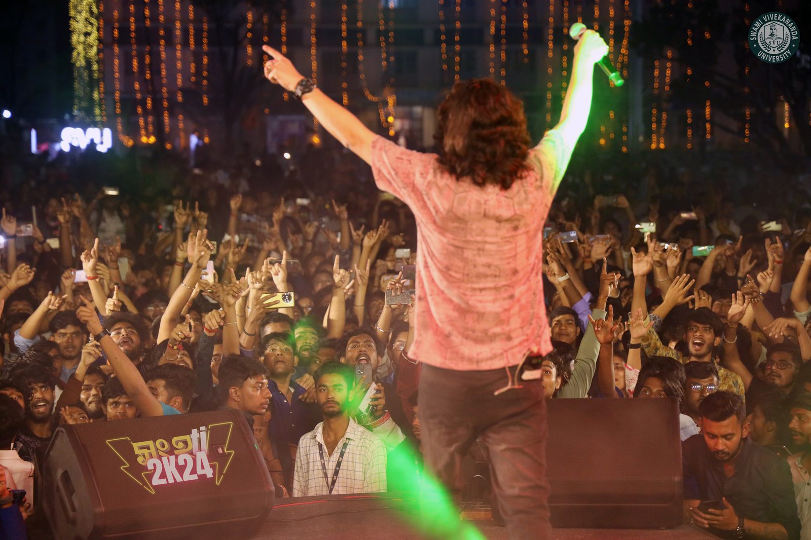 A performer seen from behind, arms spread over a night crowd of raised hands and fairy lights