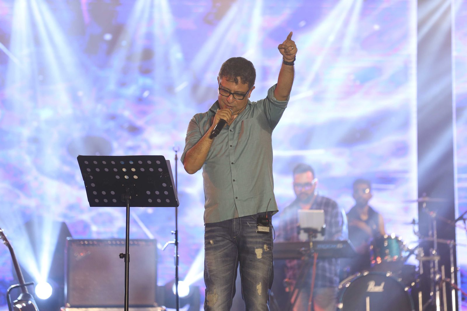 A singer with one arm up, keyboard player and drummer working behind him