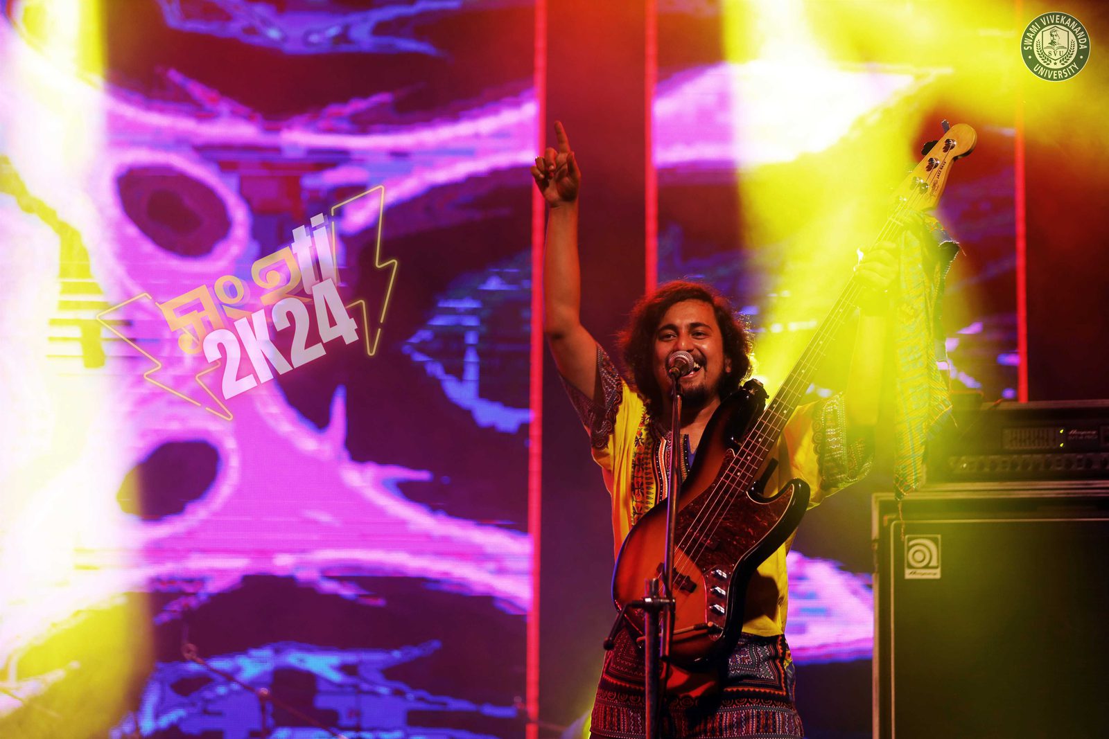 The bassist pointing skyward and grinning against a blaze of pink and yellow light